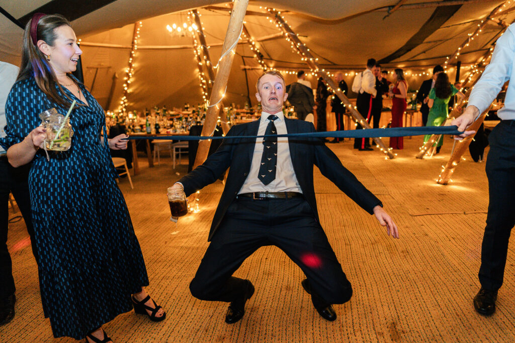 A person in a suit leans back while attempting the limbo at a festive indoor event. They hold a drink in one hand. Another person stands nearby, watching. Warm string lights and decorated tables fill the background.