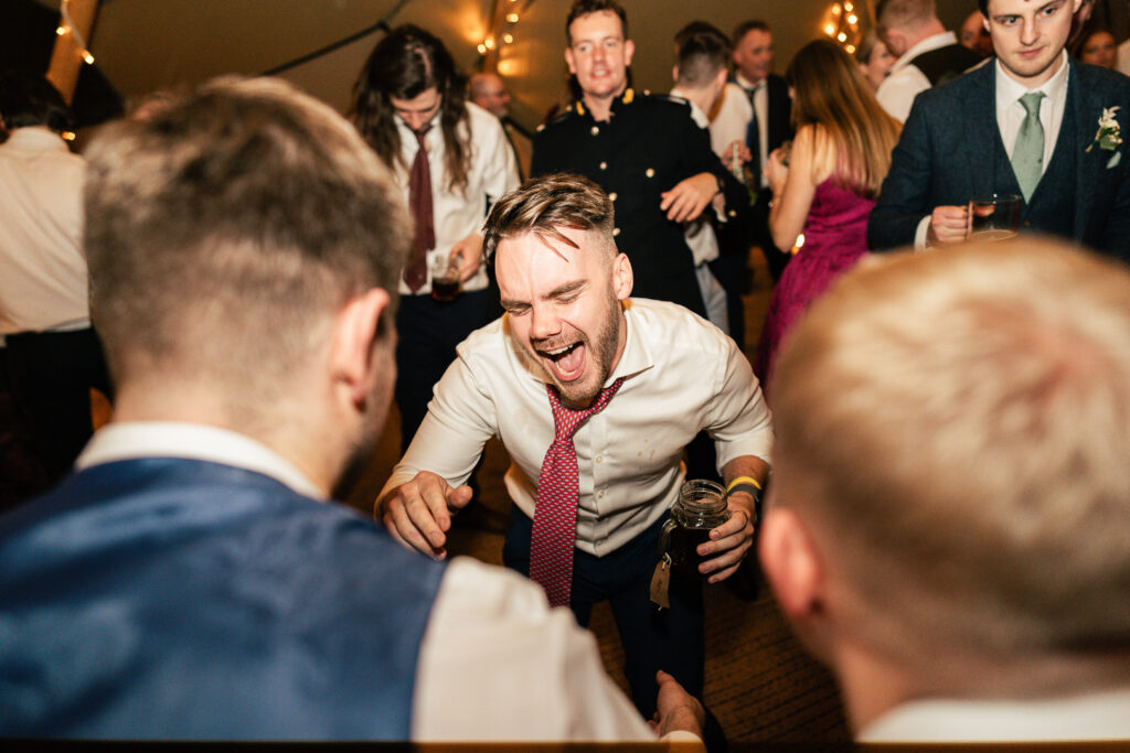 A lively party scene with a man in a white shirt and red tie energetically dancing in the center. He is surrounded by people in formal attire, some holding drinks, under warm, festive lighting.