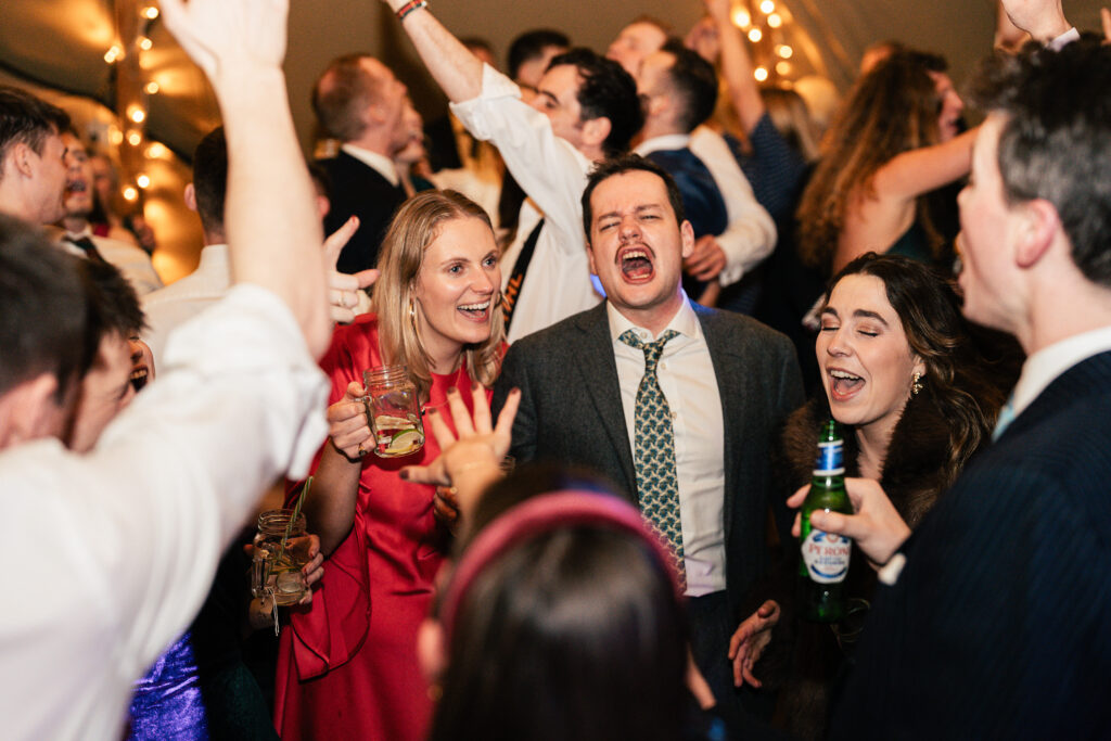 A lively group of people is dancing and singing at a party. A man in a suit and a woman in a pink dress are in the center, holding drinks. Others around them have their hands raised, with festive lights in the background.