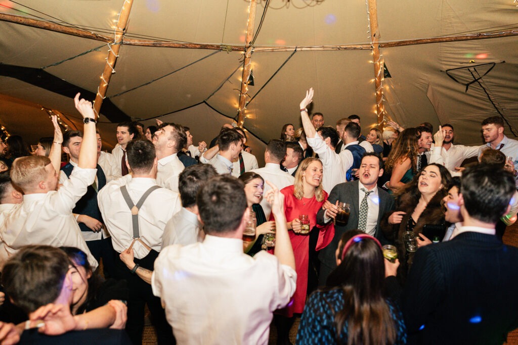 A lively party scene in a tent with people dancing, laughing, and holding drinks. String lights hang from the ceiling, adding a festive atmosphere. Guests are dressed in formal attire, enjoying the celebration.