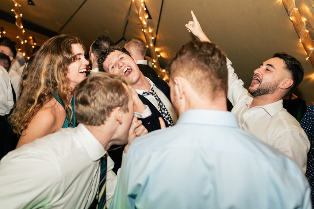 A group of people at a party are enthusiastically singing and dancing together under string lights. They are wearing formal attire, with a mix of shirts, ties, and dresses, and appear to be having a joyful time.