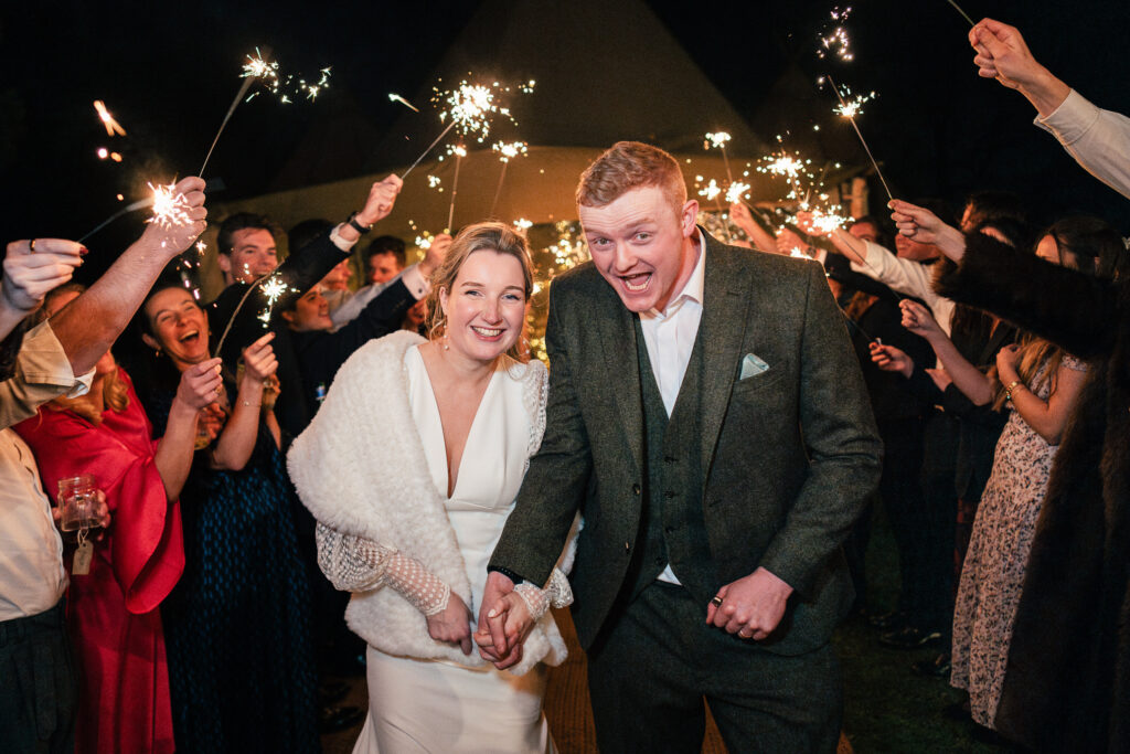 A smiling couple in wedding attire walks through a crowd holding sparklers. The scene is festive and joyful, illuminated by the sparklers and night lighting. Guests are cheering and celebrating.