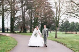 A couple walks hand in hand down a paved path lined with bare trees. The bride wears a white gown and the groom wears a gray suit. The setting is a park with green grass and a cloudy sky.