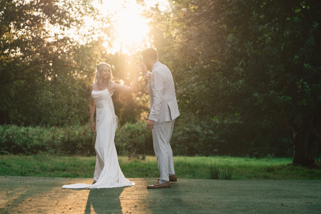 A couple stands on a grassy area during sunset. The woman, in a white dress, smiles and gently holds the man's hand. The man, in a light-colored suit, faces her. Sunlight filters through trees, creating a warm glow around them.