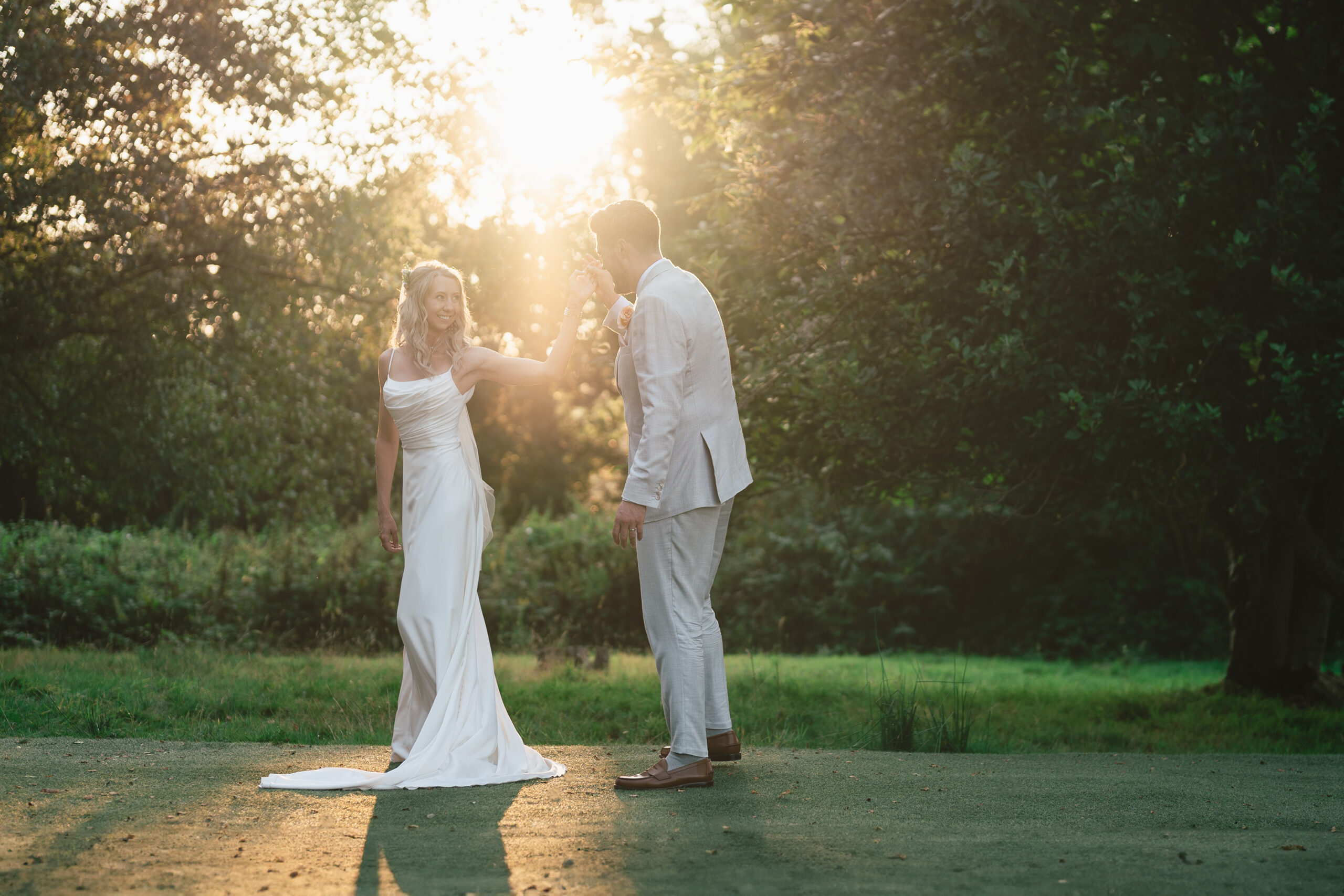 A couple in formal attire stands outdoors in the soft glow of sunset. The woman in a white dress holds the man's hand, while he wears a light-colored suit. Lush greenery surrounds them, bathed in warm, golden light.