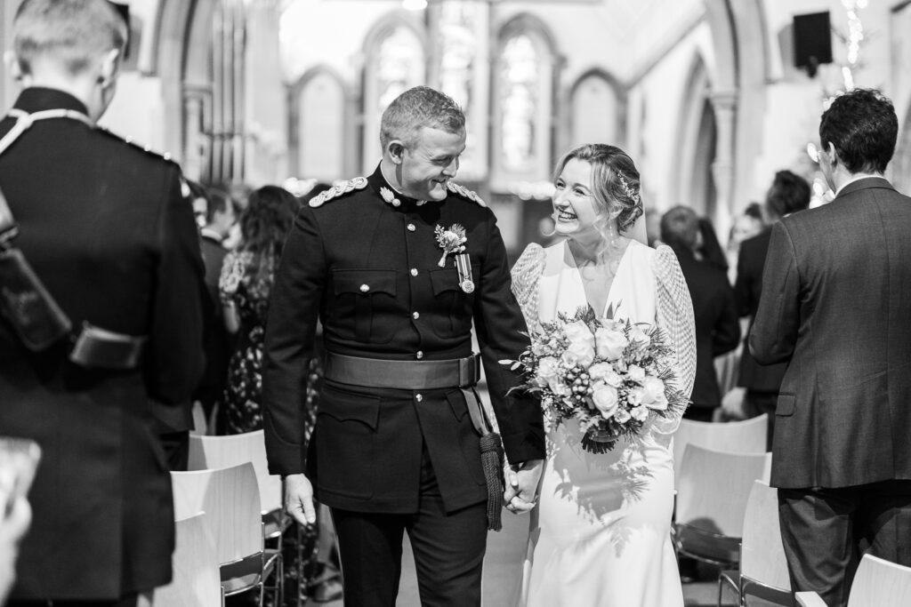 A couple smiles at each other while walking down the aisle during their wedding ceremony. The groom is in a military uniform, and the bride wears a white gown with a bouquet. Guests and church interior are visible in the background. Black and white photo.