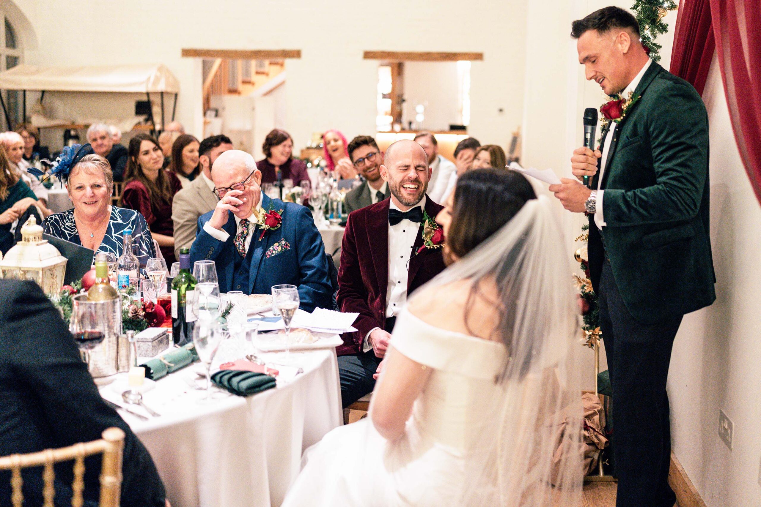 A wedding reception scene with guests seated at tables. A man stands, speaking into a microphone, smiling. A bride, in a white dress and veil, sits facing him. Decor includes rose centerpieces and glasses. Other guests are attentively watching and smiling.