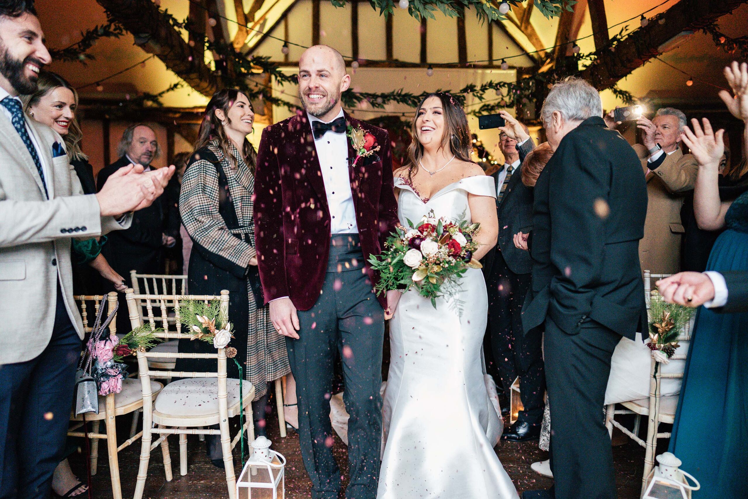 A bride and groom smile while walking down the aisle amidst clapping guests in a festive, decorated venue. The bride holds a bouquet of flowers and wears a white dress; the groom wears a burgundy jacket. Leafs and lights adorn the area.