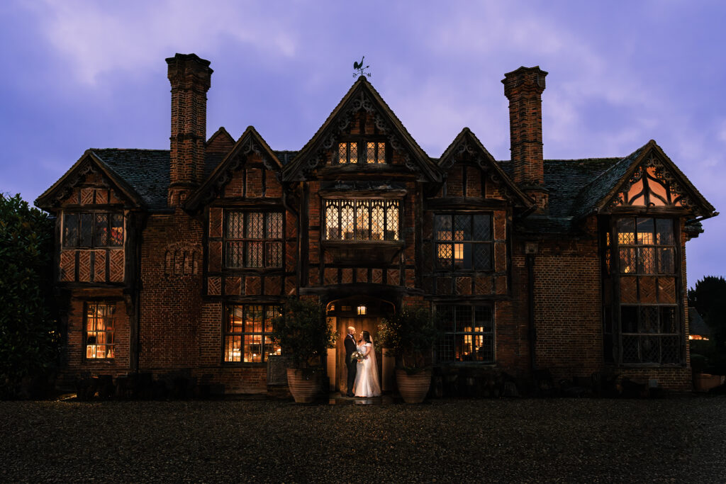 A couple in wedding attire stands in front of a large, historic brick mansion at Dinreny Court, Windsor. The building features intricate woodwork and multiple chimneys. The sky is at blue hour, adding a romantic ambiance.