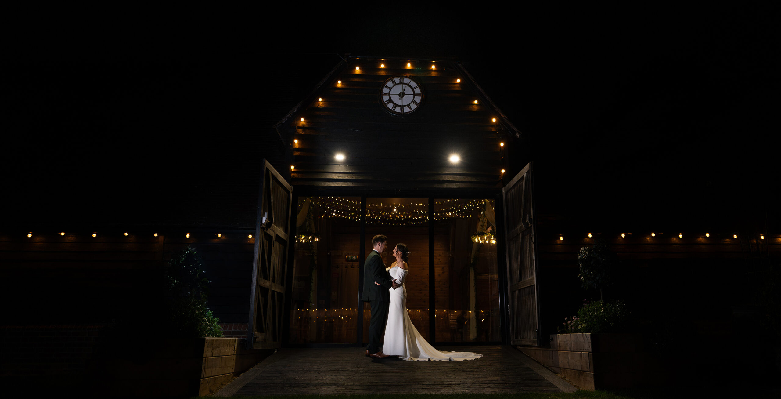 A bride and groom stand embracing in front of a barn decorated with string lights at night. The barn doors are open, and a clock is visible above. The scene is illuminated, highlighting the couple against the dark backdrop.