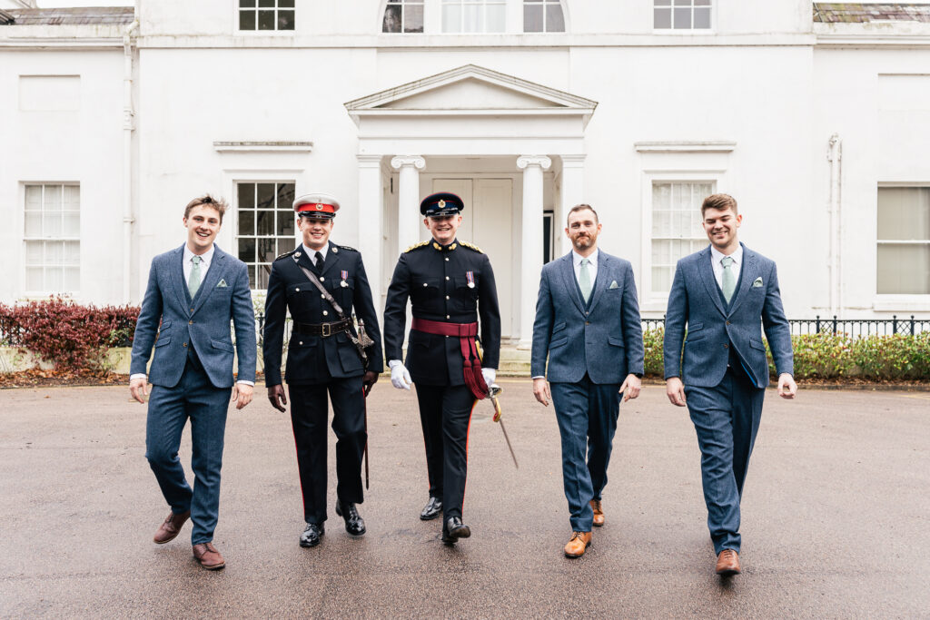 Five men walking toward the camera in front of a white building. Two are wearing military uniforms, and three are in matching blue suits with ties and pocket squares. The ground is wet, suggesting recent rain.