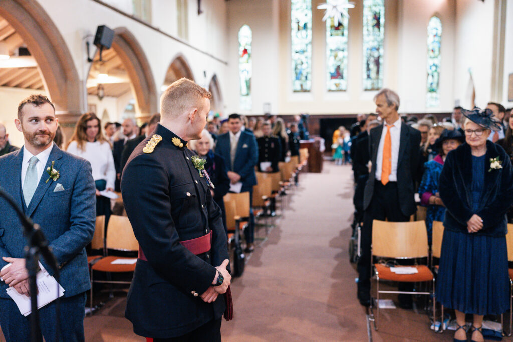 A groom in a military uniform stands at the altar, looking toward the back of a church aisle. Guests are seated and standing, with stained glass windows visible in the background. The atmosphere appears formal and anticipatory.