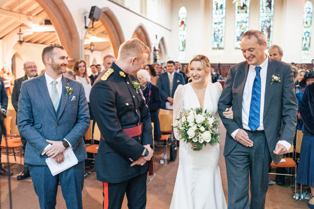 Bride in a white dress with a bouquet walks down the aisle with an older man. They are met by a man in a decorated uniform and another in a suit, smiling inside a church filled with guests.