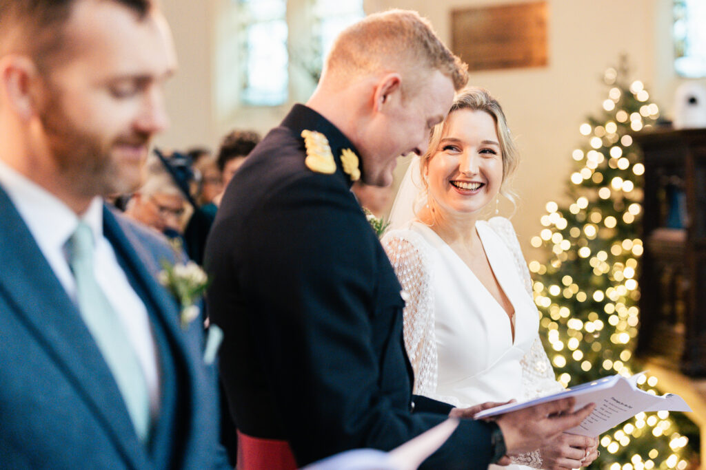 A bride and groom smile at each other during a wedding ceremony. The groom is in a military uniform and the bride wears a white dress. A man in a suit stands beside them. There are festive lights and blurred guests in the background.