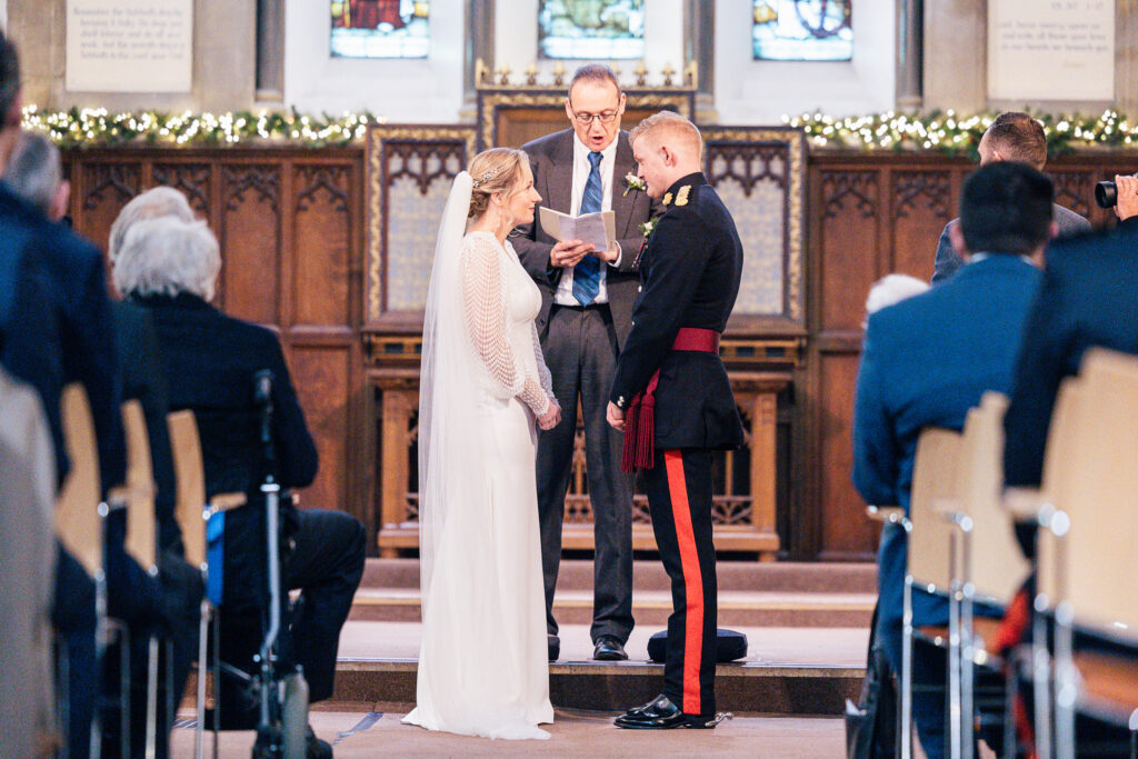 A bride and groom stand facing each other in a church during a wedding ceremony. The officiant stands behind them, reading from a paper. The bride wears a white dress, and the groom is in a formal military uniform. Guests are seated in the foreground.