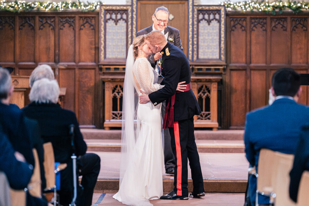 A couple shares a kiss at the altar during a wedding ceremony. The bride wears a long white dress with a veil, and the groom is in a military-style uniform. An officiant stands behind them, and guests are seated on either side in the venue.