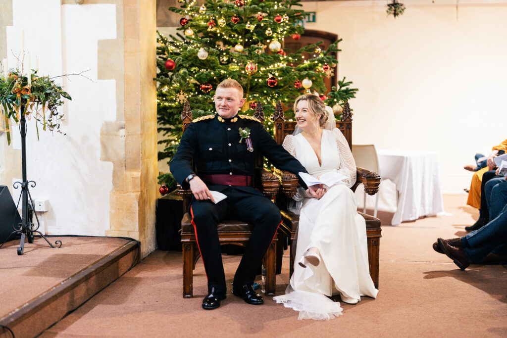 A bride and groom sit on ornate chairs, holding hands and smiling. The groom wears a formal military uniform, and the bride is in a white wedding dress. A decorated Christmas tree is in the background. The setting appears festive and celebratory.