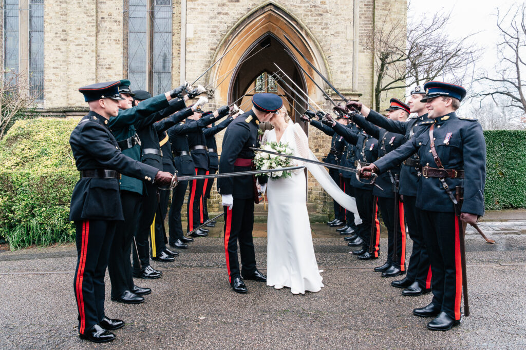 A bride and groom kiss under an archway formed by uniformed military personnel holding swords aloft. The bride is in a white dress, and the groom is in a military uniform. They are outside, standing in front of a historic stone building.