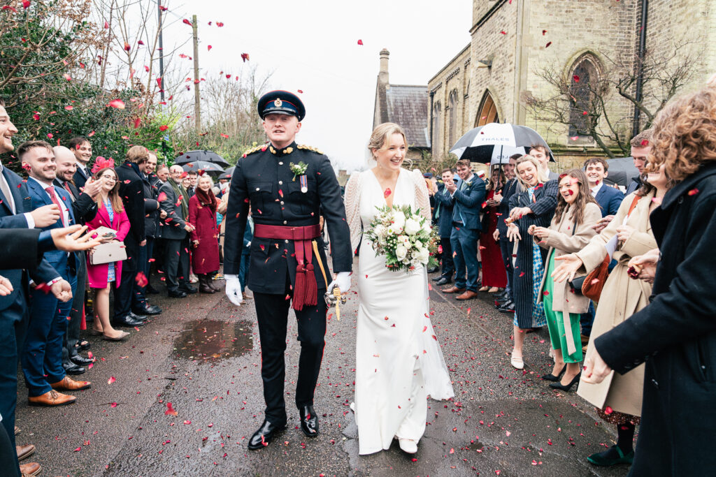 A bride and groom walk hand in hand outside a church while guests throw rose petals. The bride wears a white gown and holds a bouquet, and the groom is in a formal military uniform. Guests surround them, celebrating with smiles and umbrellas.