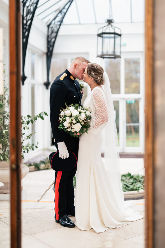 A bride and groom share a kiss in a bright, elegant room. The groom is in a formal military uniform, holding a hat and gloves, while the bride wears a long white dress with a veil. They are surrounded by greenery and natural light.