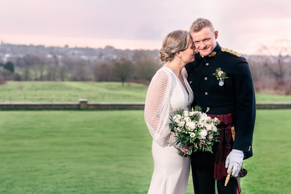 A bride and groom stand close together on a lush green lawn. The bride, in a white dress holding a bouquet, leans towards the groom. The groom wears a formal military dress uniform. A soft, cloudy sky and distant landscape are in the background.