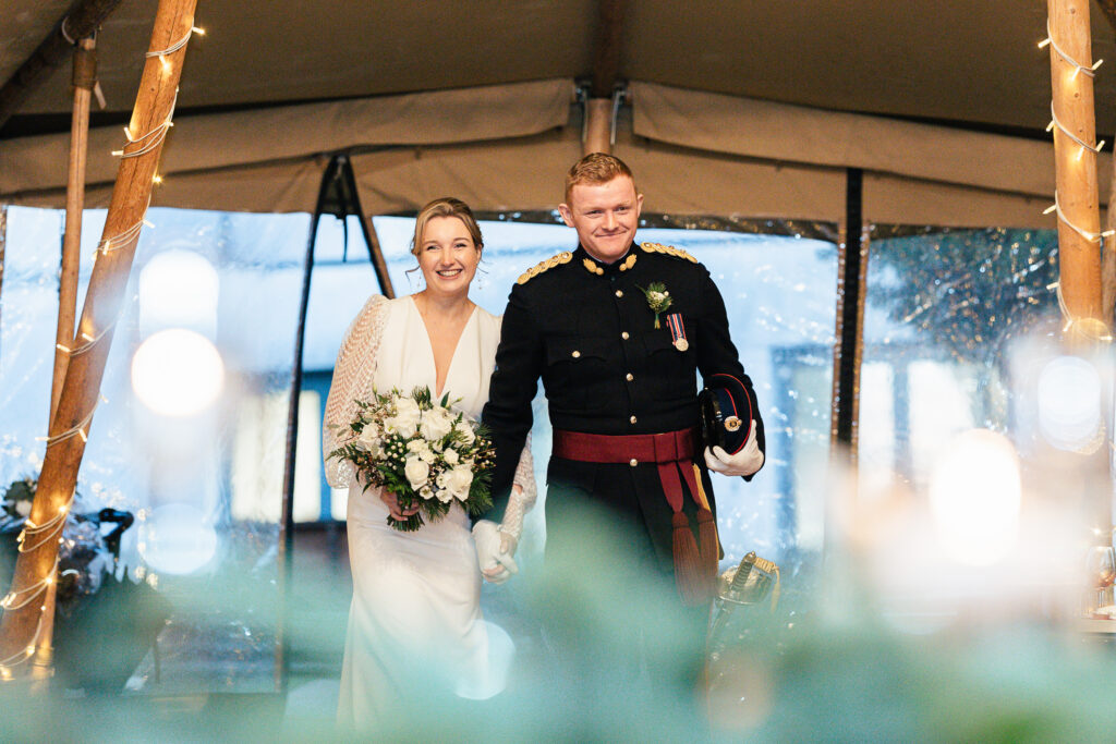 A couple is smiling and holding hands at a wedding. The bride is in a white dress holding a bouquet, and the groom is in a dark military uniform with medals. They are standing under a tent adorned with lights.