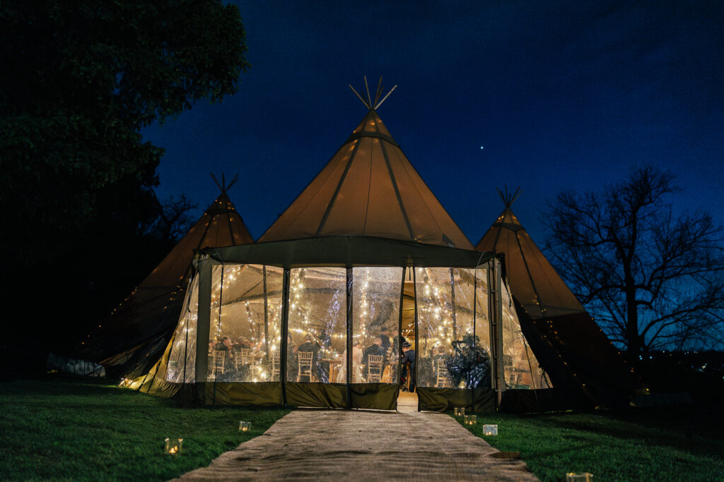 A large outdoor tent illuminated with string lights is set up at night. The tent has a pointed top, and there's a star visible in the clear dark sky. The entrance is framed with transparent panels, revealing a warmly lit interior.