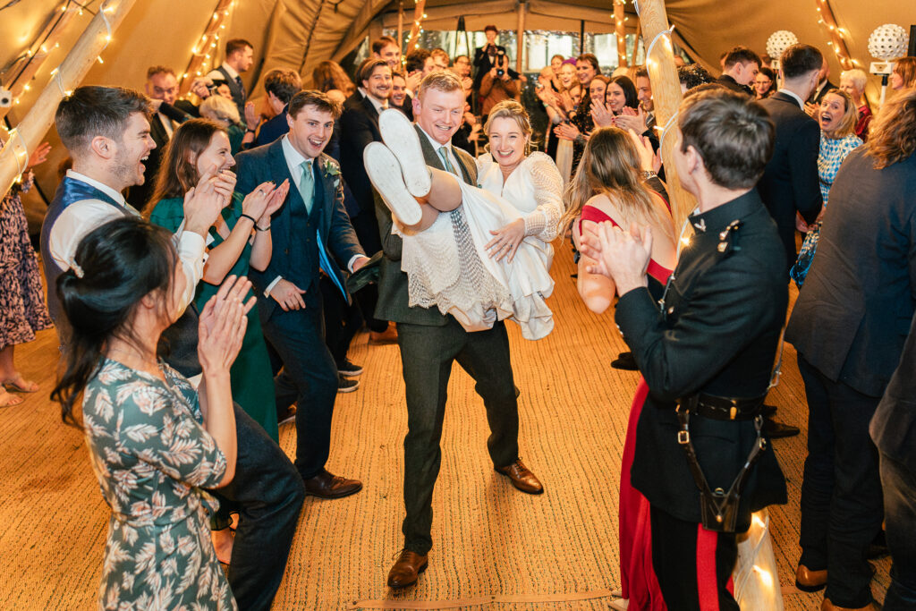 A couple is laughing joyfully in the center of a lively dance floor; the groom is carrying the bride. They are surrounded by cheering guests in festive attire inside a warmly lit tent.