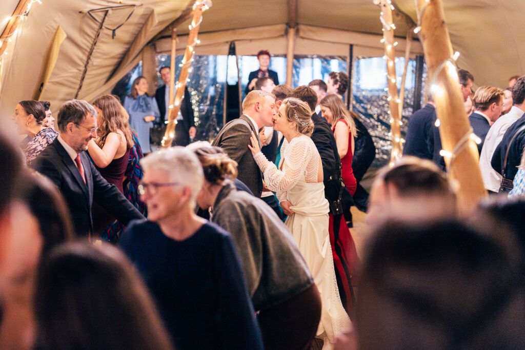 A couple kisses surrounded by people dancing at a festive event inside a tent decorated with string lights. Many guests are dressed in formal attire, and the atmosphere appears lively and celebratory.