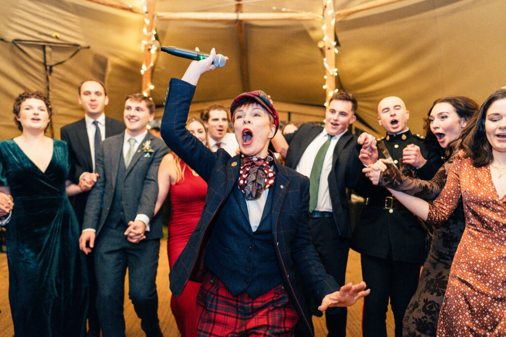 A lively group of people in formal attire, gathered under a warmly lit tent. One person, dressed in a plaid suit and cap, energetically holds a microphone, appearing to sing or speak, as others around them smile and join in the excitement.