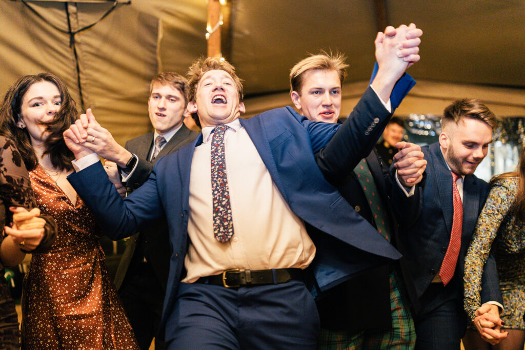 A group of people in formal attire are enthusiastically dancing. A man in a blue suit and patterned tie is in the center, raising his arms and cheering. Others around him, both men and women, are smiling and joining in the celebration.