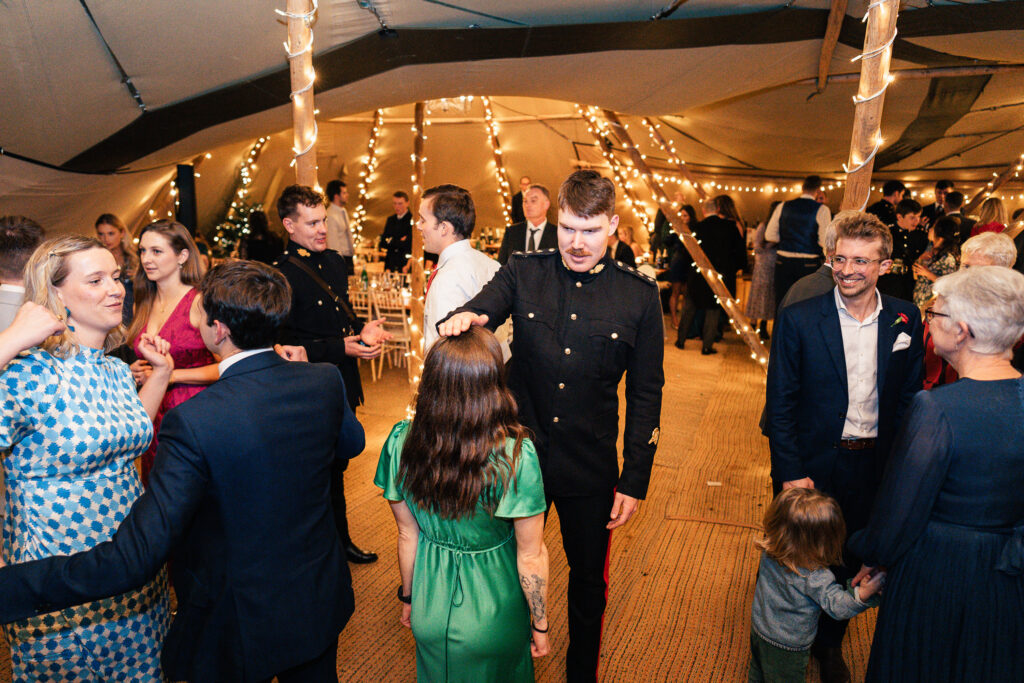 People dancing at an indoor event with string lights decorating the area. A man in a black suit interacts with a woman in a green dress. Several others are mingling and dancing around them in a festive atmosphere.