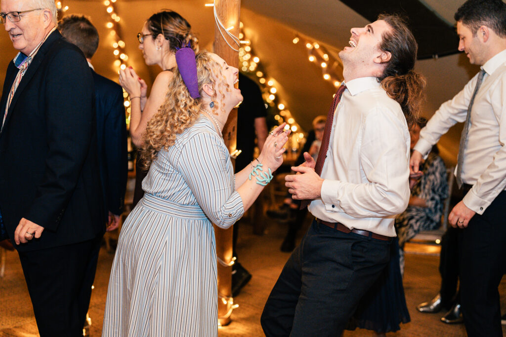 People are dancing and laughing at a festive indoor event. The room is decorated with string lights. A man with long hair and a woman with curly hair stand out in the center, both looking joyful. Others are dancing around them.