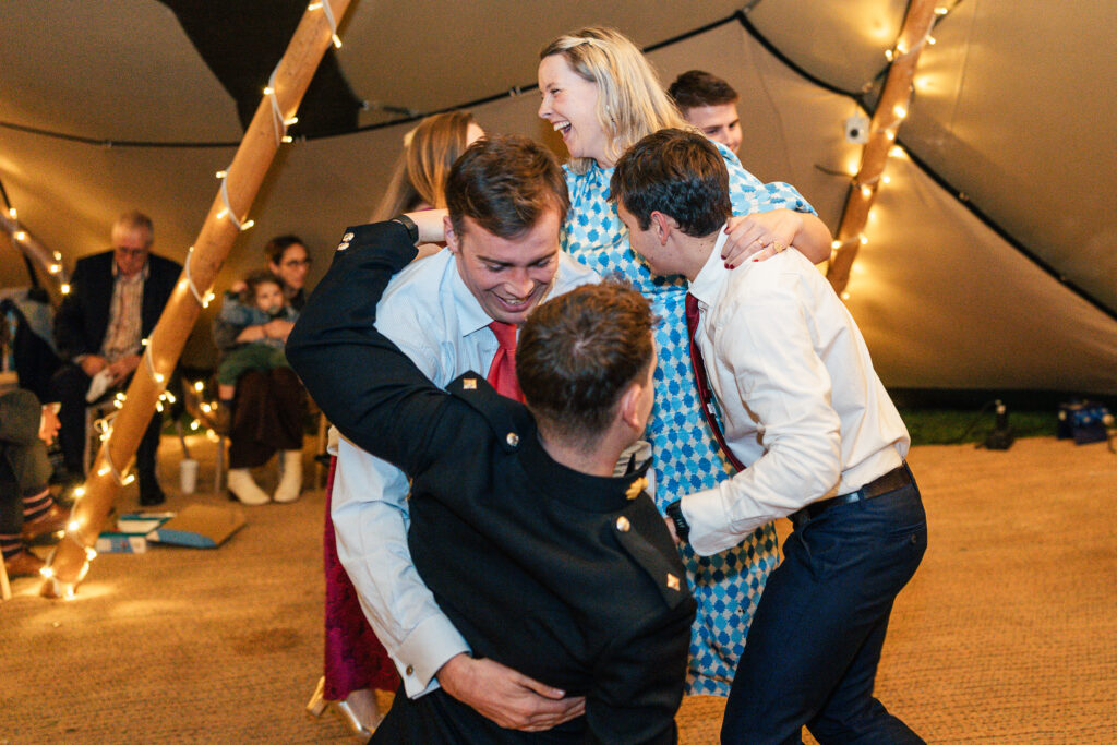 A joyful group of people dance energetically inside a warmly lit, tented area. A man in a kilt swings another man in a light blue shirt around, while others laugh and move around them. String lights wrap around the tent poles.