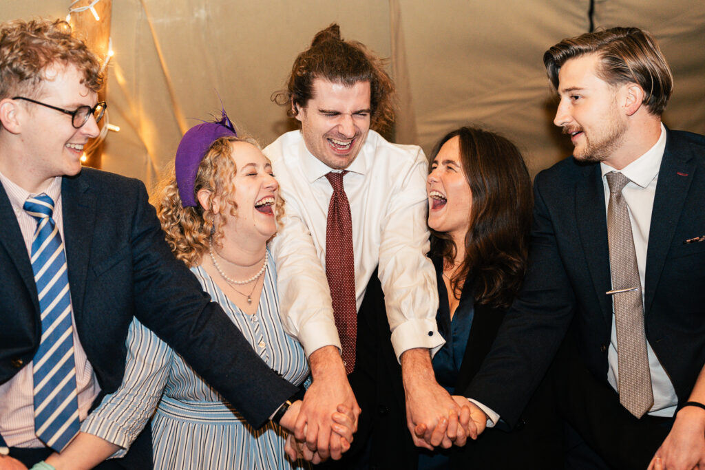 Five people dressed formally are standing in a circle, holding hands and laughing. They appear to be celebrating at an indoor event with festive lighting. The mood is joyful and lively.