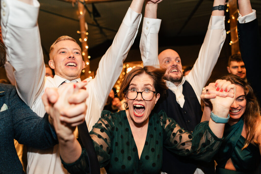 A group of people dressed in formal attire enthusiastically raise their hands. The woman in the foreground has glasses and a green polka-dotted dress. The setting appears festive with string lights in the background.