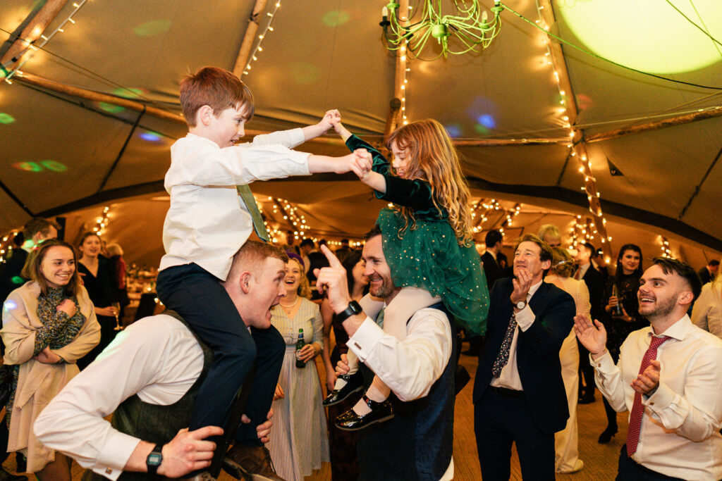 A joyful indoor party scene with two men holding kids on their shoulders. The kids are playing and holding hands above. Surrounding them, people are smiling, clapping, and enjoying the festive atmosphere with string lights overhead.