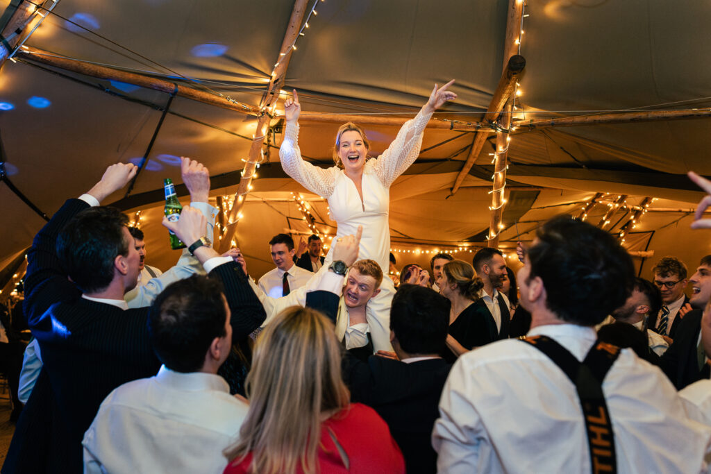 A woman in a white outfit is joyfully lifted in the air by a crowd at a lively indoor celebration. The room is decorated with strings of lights, and people around her are cheering and raising their hands in excitement.