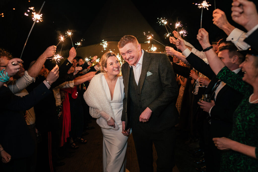 A bride and groom walk through a crowd of people holding sparklers at night. The bride wears a white dress with a shawl, and the groom is in a dark suit. Both are smiling as guests cheer them on, creating a festive atmosphere.