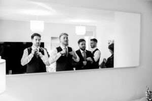 A black and white photo of four men wearing suits and adjusting their ties in front of a large wall mirror. A woman stands nearby. Two ceiling lights are visible above them.