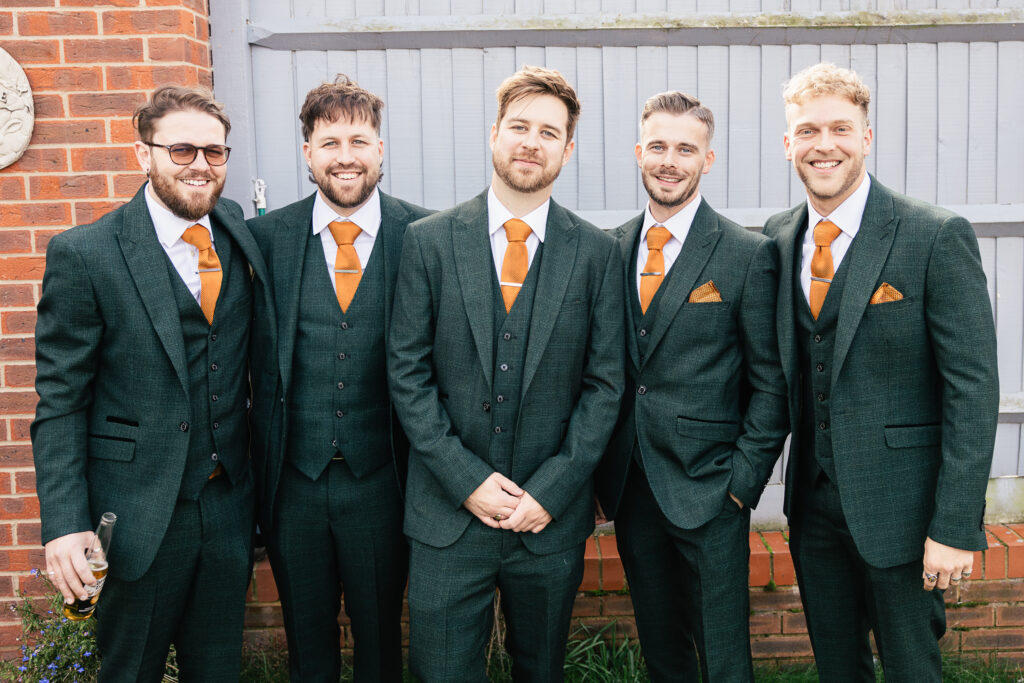 Five men in matching dark green suits with orange ties stand side by side, smiling. They are in front of a brick wall and a wooden gate. One man holds a drink in his hand. The background includes small plants.