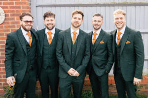 Five men in matching dark green suits with orange ties stand side by side, smiling. They are in front of a brick wall and a wooden gate. One man holds a drink in his hand. The background includes small plants.