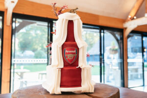 A multi-tiered wedding cake designed to resemble an Arsenal football jersey is adorned with cascading white fondant drapes and floral accents. It stands on a wooden table in a bright room with large windows overlooking greenery.