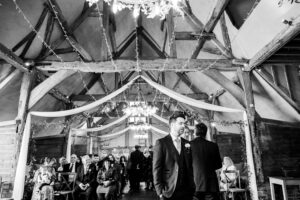 A black and white photo of a rustic wedding ceremony inside a barn with wooden beams. Guests are seated, and a man in a suit stands at the front. Draped fabric and string lights decorate the ceiling.