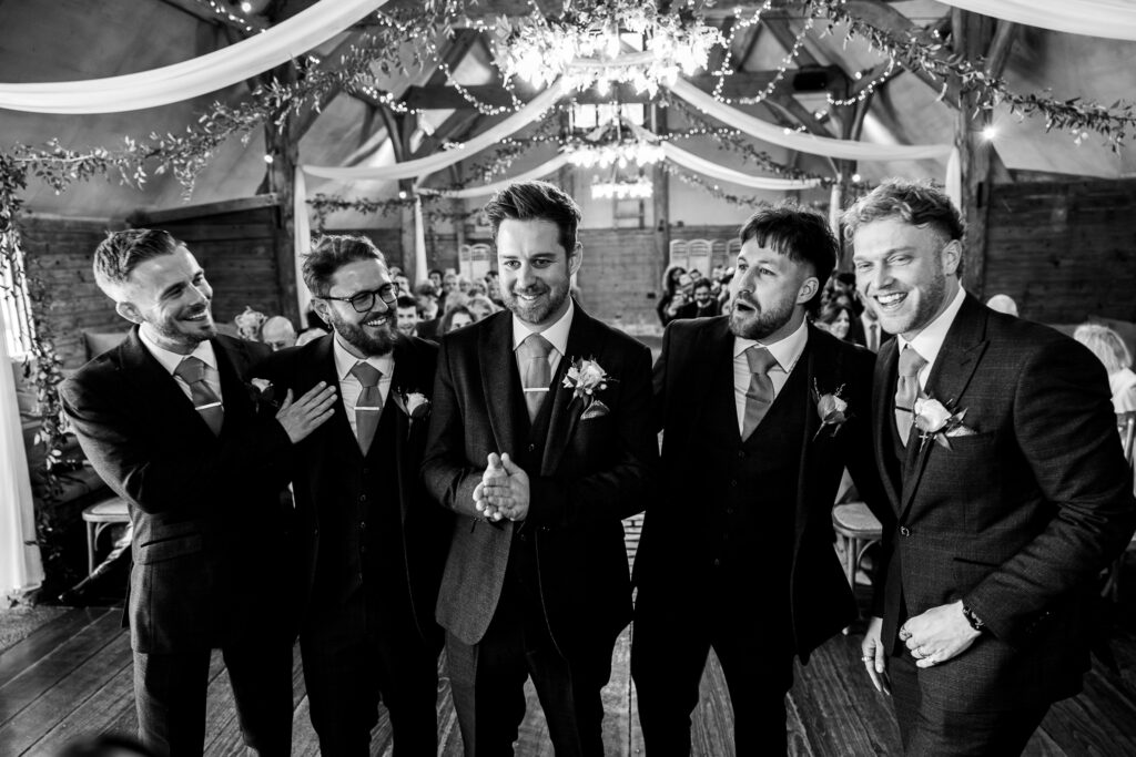 Five men in suits, wearing boutonnieres, stand together smiling and chatting in a decorated indoor venue. The ceiling is adorned with drapes and string lights, creating a festive ambiance. The scene captures a joyful, celebratory moment.