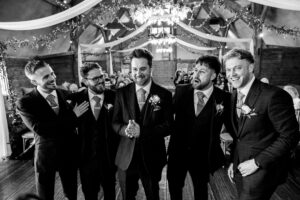 Five men in suits, wearing boutonnieres, stand together smiling and chatting in a decorated indoor venue. The ceiling is adorned with drapes and string lights, creating a festive ambiance. The scene captures a joyful, celebratory moment.