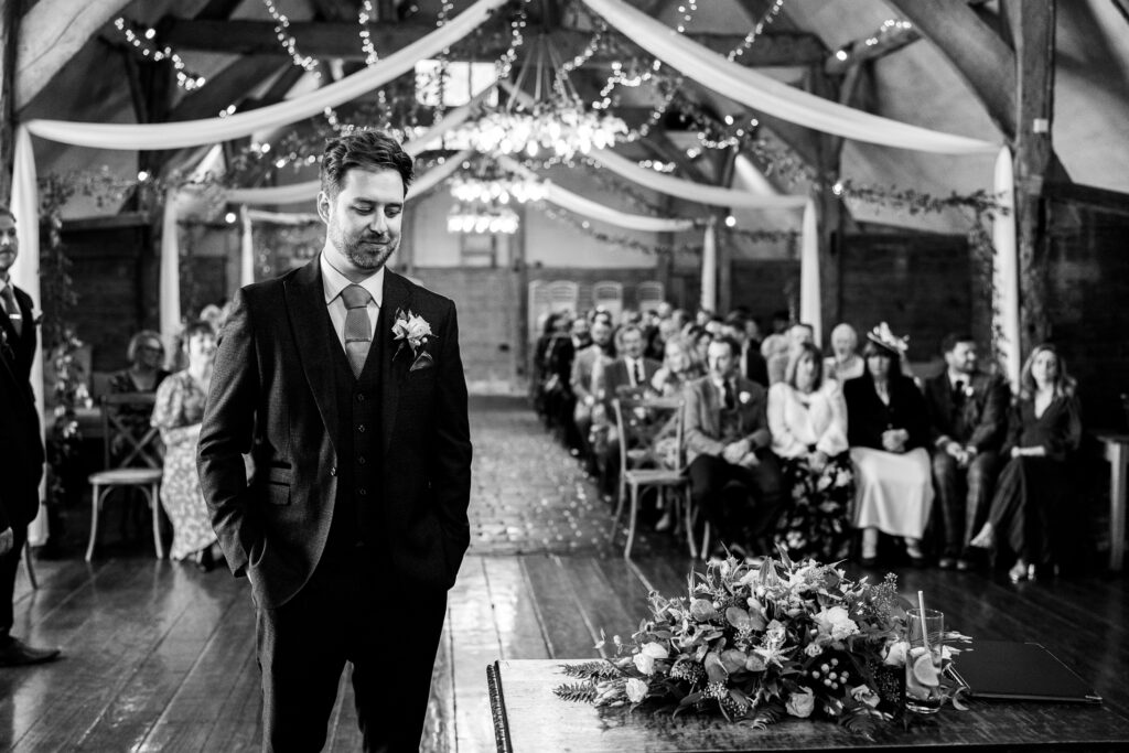 A man in a suit stands at the altar of a decorated wedding venue, looking downward with hands in pockets. Guests are seated in rows behind him, with draped fabric and lights hanging from the ceiling. A floral arrangement is on the table in front of him. Black and white.