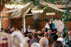 A wedding ceremony in a rustic, wood-paneled venue. The bride and groom stand at the altar under a decorated archway with greenery. Guests are seated, watching attentively. Soft, ambient lighting creates a warm atmosphere.