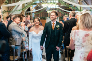 A bride and groom smile and walk hand in hand down the aisle in a rustic venue. Guests on either side clap and celebrate. The bride wears an off-shoulder dress, and the groom wears a dark suit and orange tie. The venue is decorated with greenery and draped fabric.