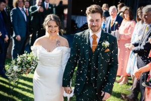 A bride in a white dress holding a bouquet and a groom in a dark suit with an orange tie smile as they walk hand in hand. Confetti is being thrown by a crowd of well-wishers around them on a sunny day.